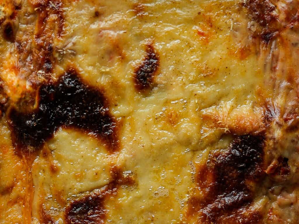 Close-up of a golden-brown, baked cheesy surface with some dark, crispy spots. The texture appears melted and slightly bubbly, suggesting a dish like lasagna or a casserole.